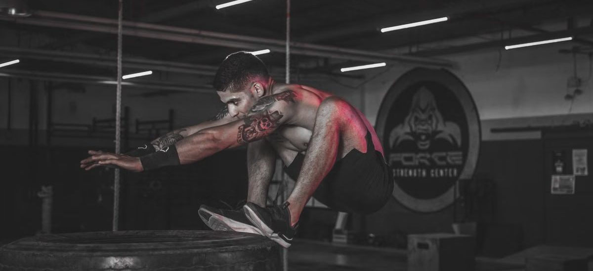 Tattooed man jumping onto large tire indoors at a gym.