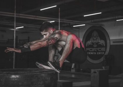Tattooed man jumping onto large tire indoors at a gym.